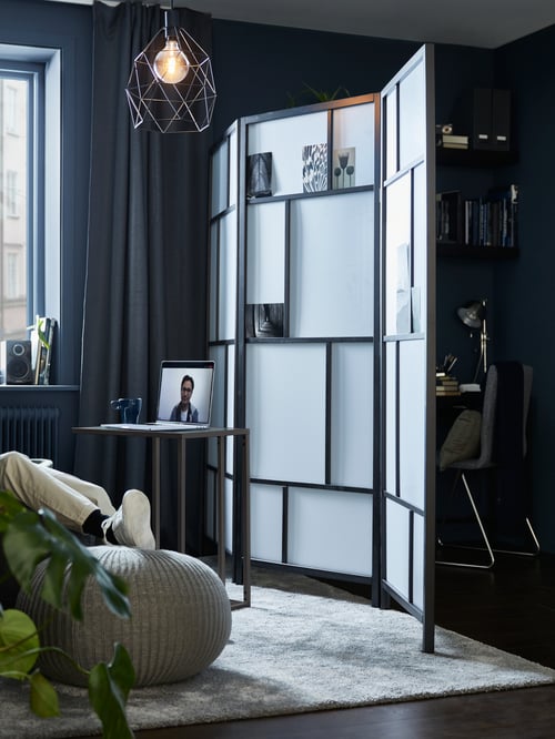 Un salon lugubre dans lequel on retrouve un paravent rectangulaire en bois noir et blanc, des rideaux gris jusqu’au sol, un pouf gris et un tapis gris.