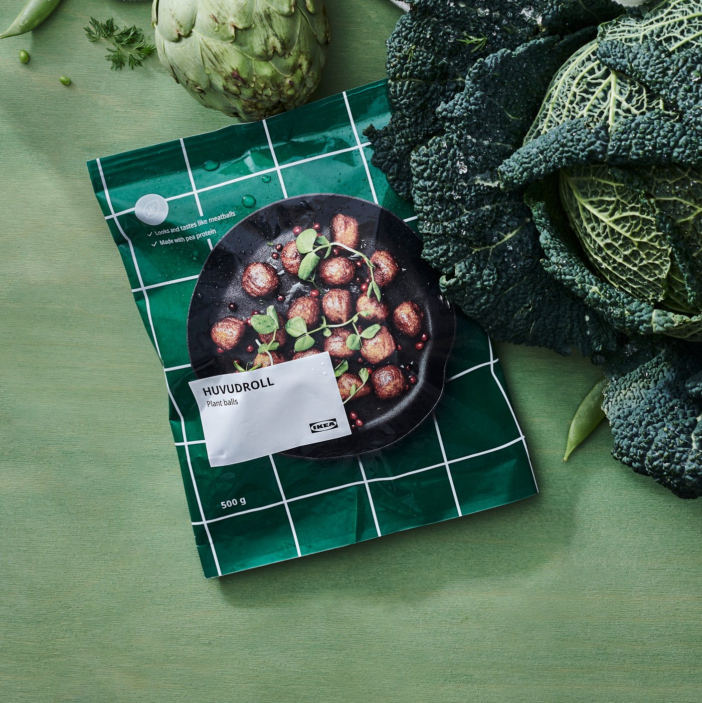 Pakiranje proizvoda HUVUDROLL s biljnim okruglicama na zelenoj podlozi, okruženo svježim povrćem poput kelja i artičoke.