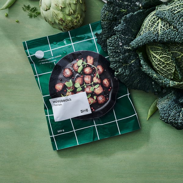 Eine Packung pflanzliche Proteinbällchen liegt umgeben von einem Kopf Wirsing und einer Artischocke auf einer grünen Fläche.
