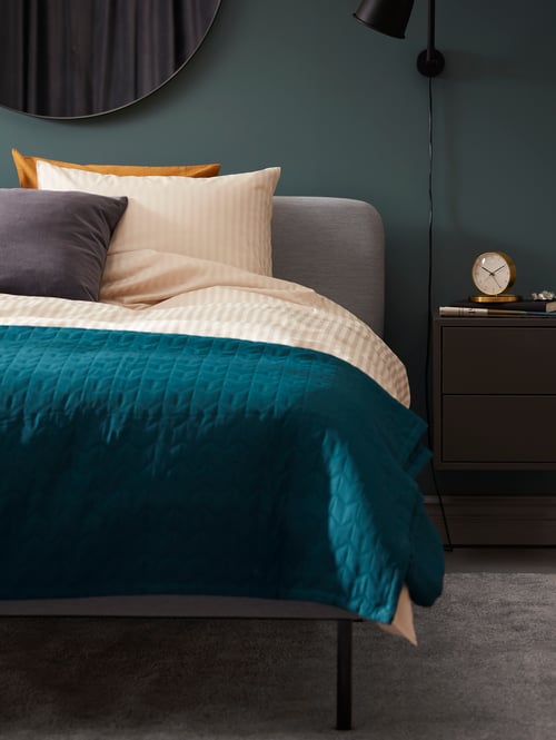 A bed with NATTJASMIN duvet cover and pillowcase, cushion and green KÖLAX bedspread and alarm clock on bedside table.