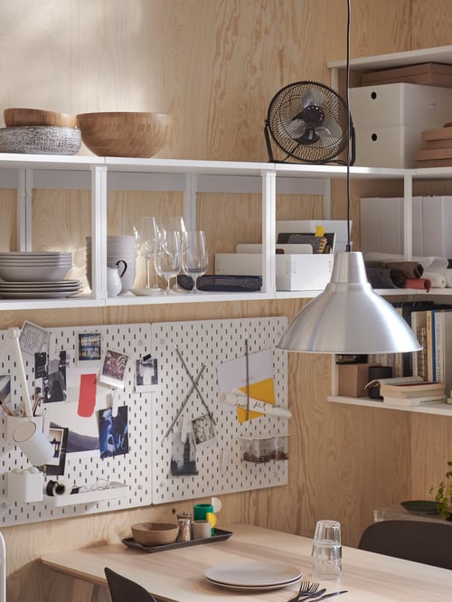 Kitchen with shelving units, dining table, cutlery, chairs, plates, glasses, bowls, pegboard, pendant lamp, box with lid.