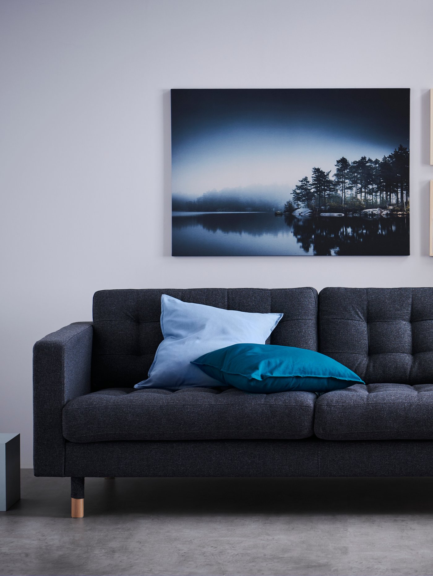 Salón con un sofá gris de tres plazas, fundas de cojín en azul y azul claro y una imagen de un lago y un bosque en la pared.
