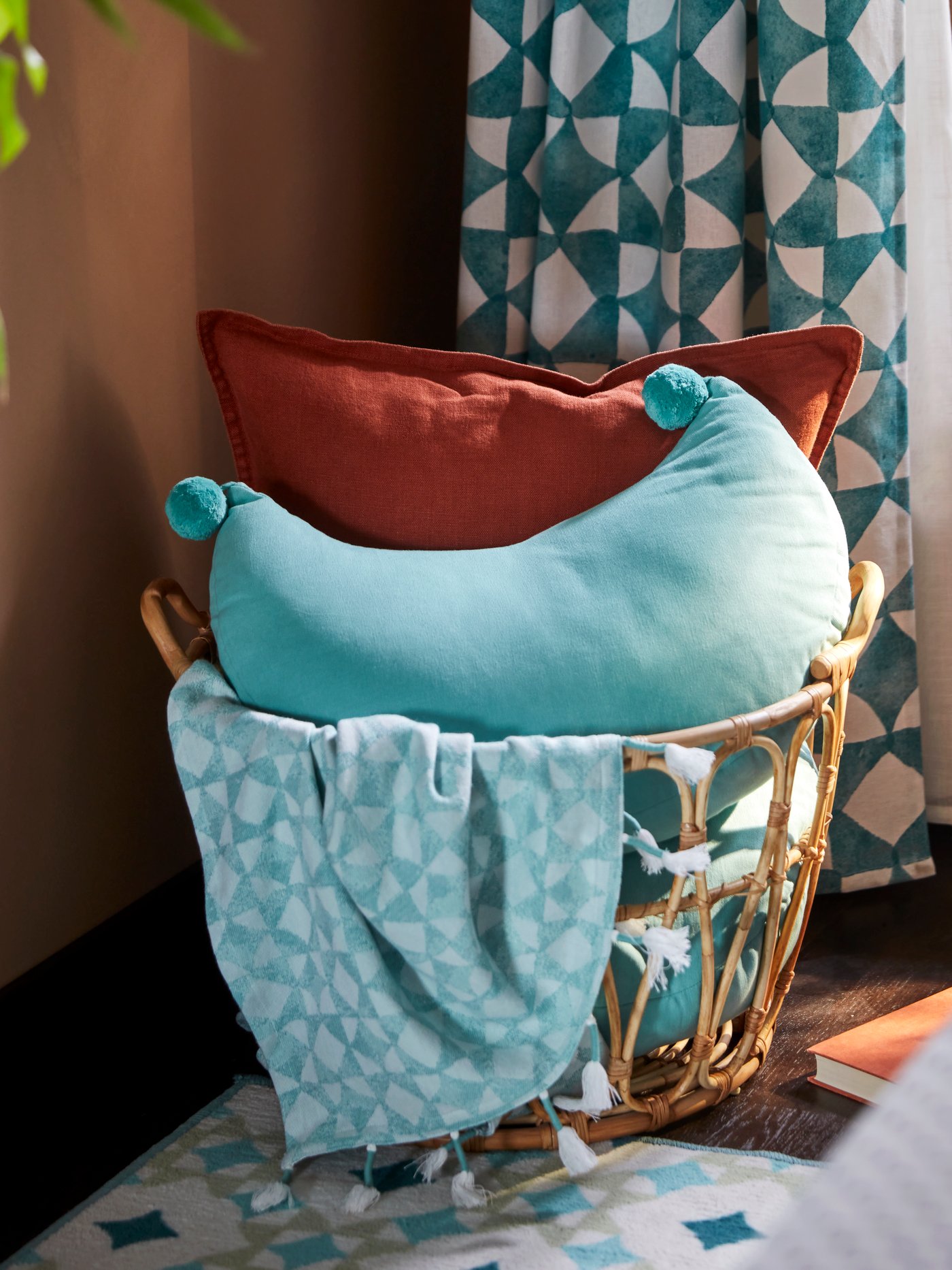 A blue GOKVÄLLÅ throw and moon-shaped cushion in a rattan basket in front of a blue patternd GOKVÄLLÅ  curtain.