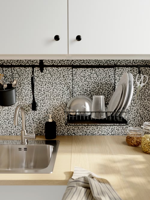 A beige kitchen worktop with an inset sink and a rail on the wall behind it with utensils and a dish drainer hanging on it.