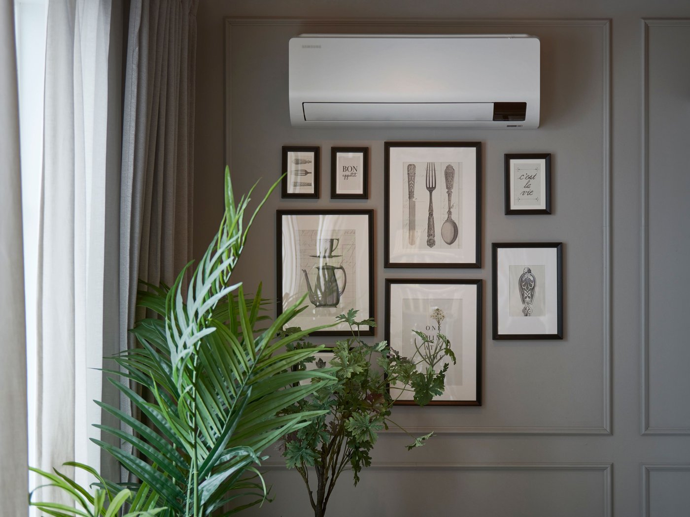 Vägg med flera inramade svartvita illustrationer av köksredskap och växter, bredvid en grön krukväxt och under en vit värrmepump. Ljuset kommer från ett fönster med ljusa gardiner till vänster.