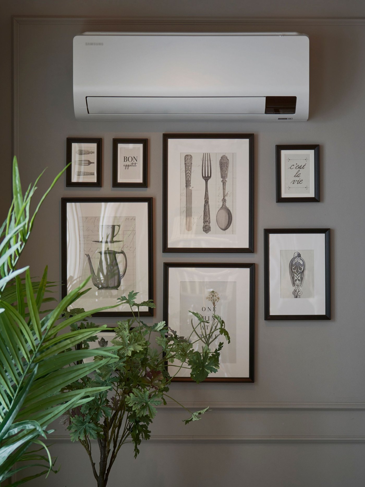 Vägg med flera inramade svartvita illustrationer av köksredskap och växter, bredvid en grön krukväxt och under en vit värrmepump. Ljuset kommer från ett fönster med ljusa gardiner till vänster.
