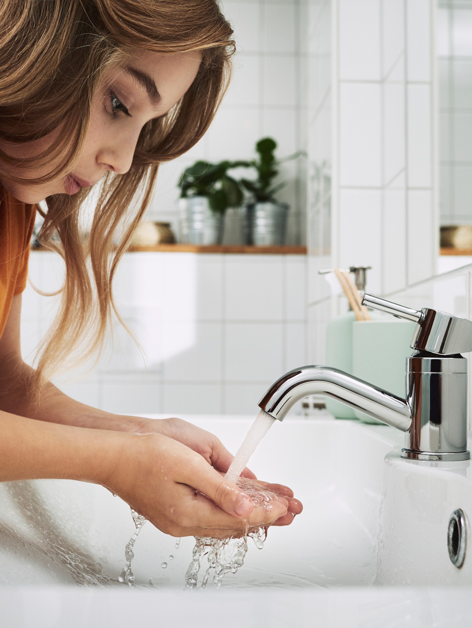 Malo dete duge smeđe kose stoji u kupatilu s belim pločicama i pere ruke u umivaoniku.