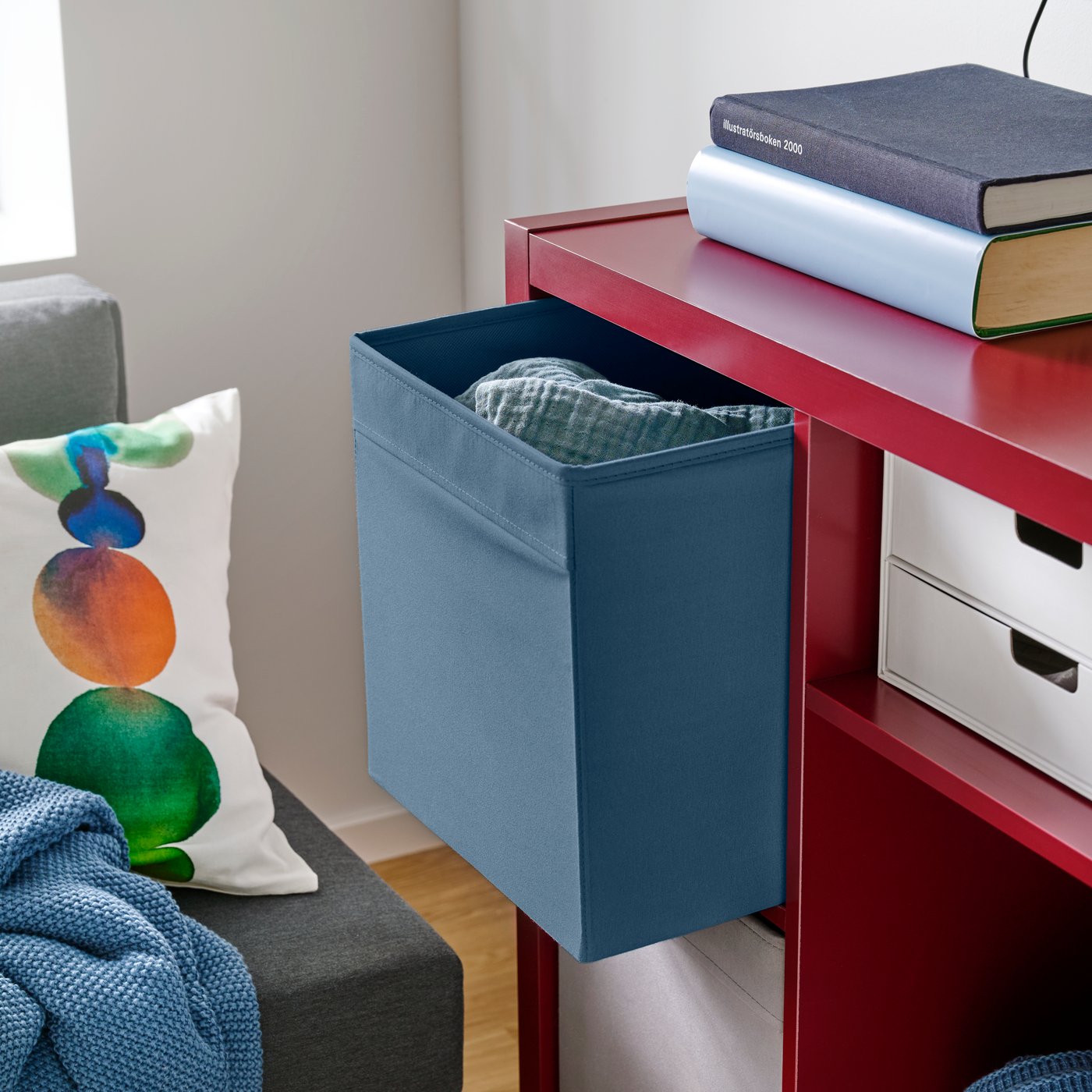 A living room with a blue DRÖNA box placed on a red shelf, beside a green and white patterned cushion.