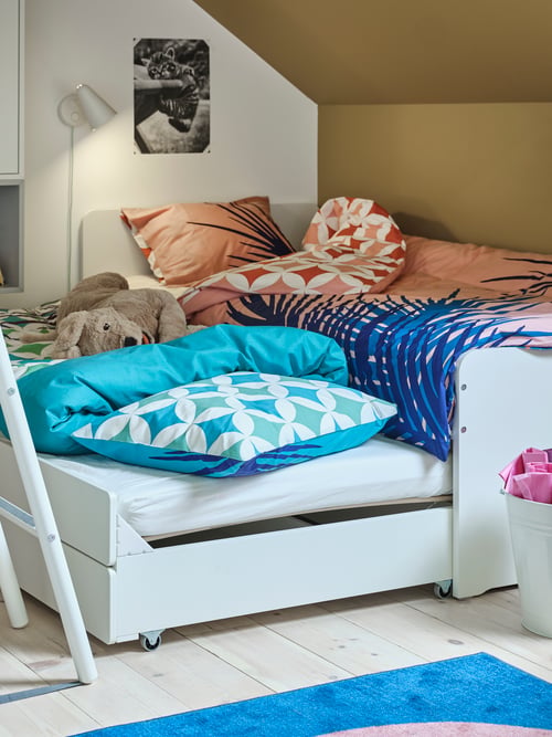A white SLÄKT bed frame with underbed and storage is extended to make two beds that have vibrant bedding and a soft toy dog.