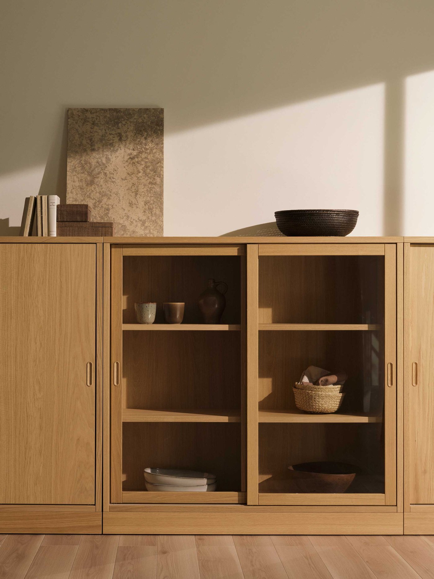 A beautifully organised wooden storage unit, perfect for keeping your living space tidy and stylish. This versatile piece combines closed cabinets for discreet storage with glass-fronted sections, allowing you to display your favourite items. Inside the glass sections, you'll spot neatly stacked plates, a charming woven basket, and some lovely ceramic cups and a vase, all ready to be used or admired. On top, a decorative dark woven bowl adds a touch of texture, alongside some books and an abstract art piece, creating a personal and inviting feel. The warm wooden tones and clean lines make this a wonderful addition to any home, helping you create a space that’s both functional and full of character.