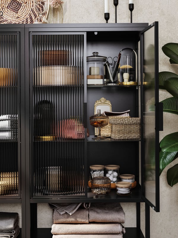 A black MOSSJÖN glass door cabinet has one door open and there are glass jars, boxes and containers on the shelves inside.