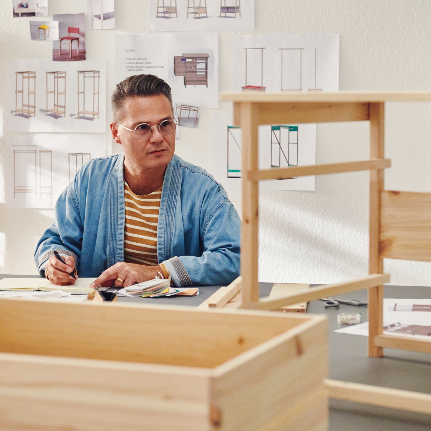 En designer i et arbeidsrom med skisser på veggen, ved siden av en KRONÖREN kommode med 2 skuffer.