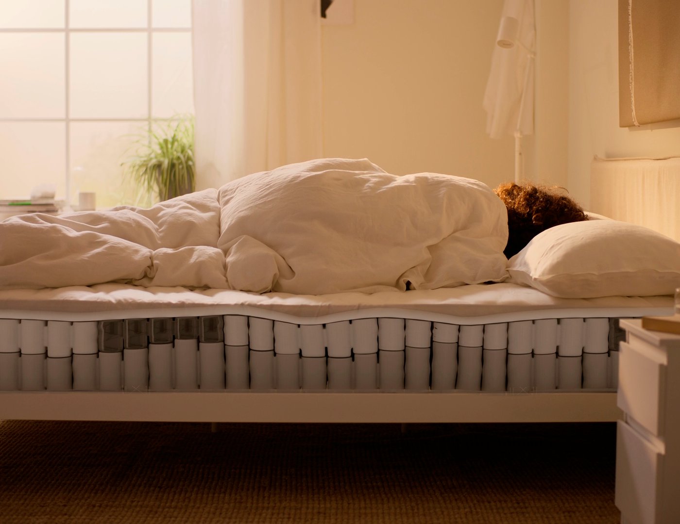 En person ligger nedbäddad i en säng i ett ljust sovrum, med madrassens fjädrar synliga genom en genomskärning i sängramen.