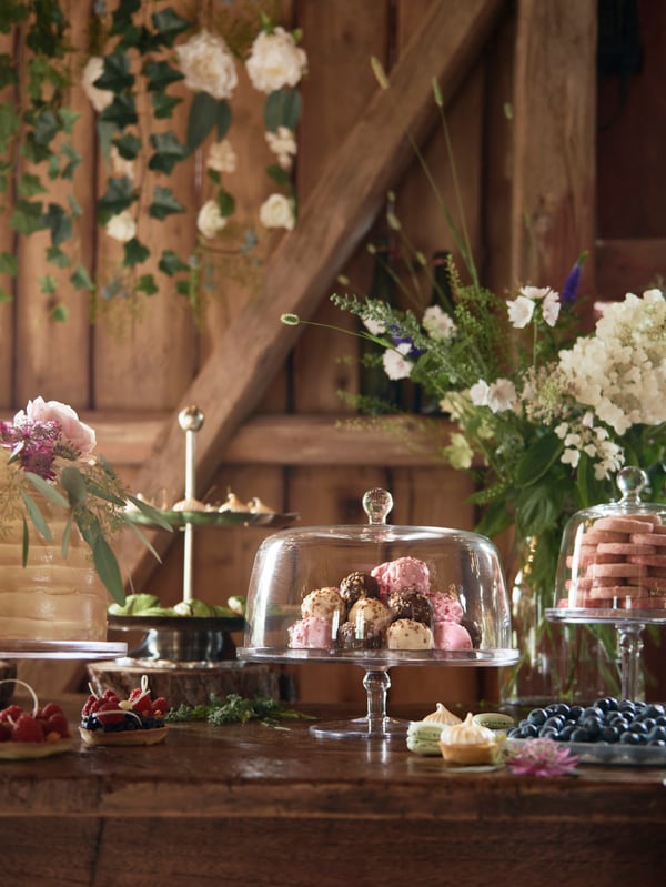 En una fiesta, tres fuentes para servir INBJUDEN llenas de dulces sobre una mesa de madera con flores y otras pastas.