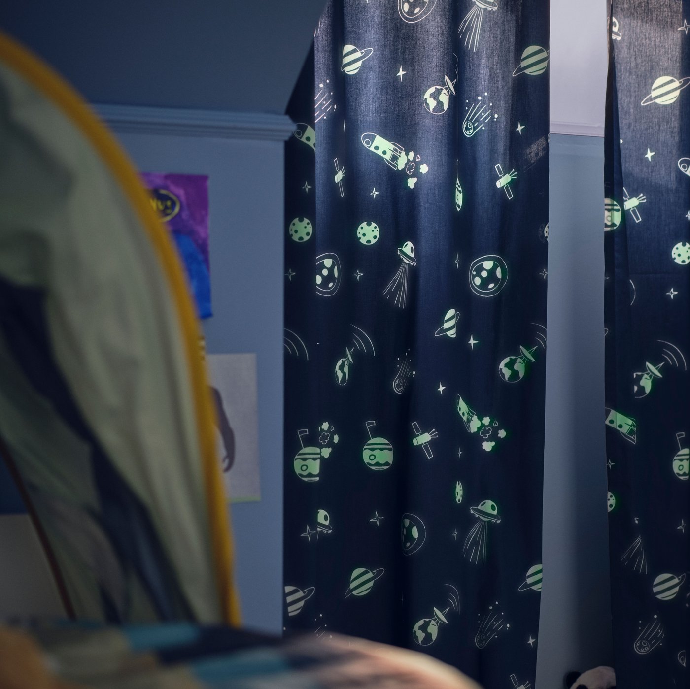 Potemnělý dětský pokoj laděný do modré barvy se zataženým závěsem AFTONSPARV, na kterém ve tmě září vesmírné motivy.