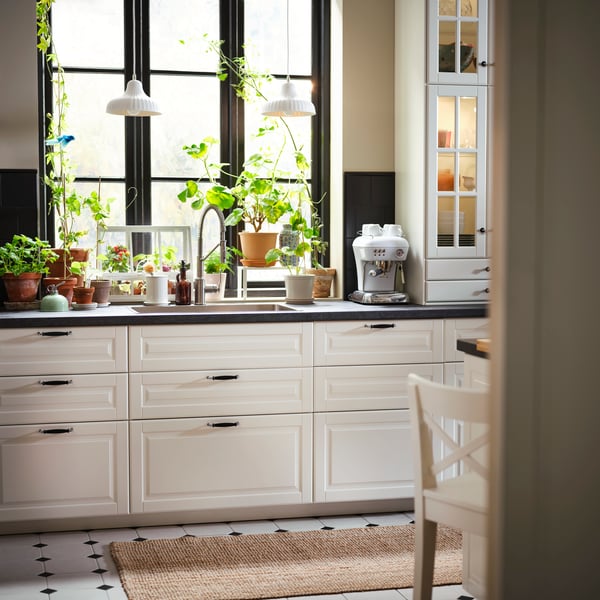 A traditional kitchen featuring BODBYN kitchen fronts in off-white, large windows with plants, and a coffee machine.