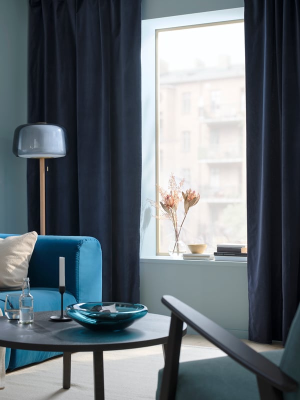 A cozy living room with light blue walls, a dark blue sofa with beige and brown cushions, a round black coffee table, and dark blue SANELA curtains framing a window. A floor lamp stands beside the sofa, and decorative items sit on the windowsill.