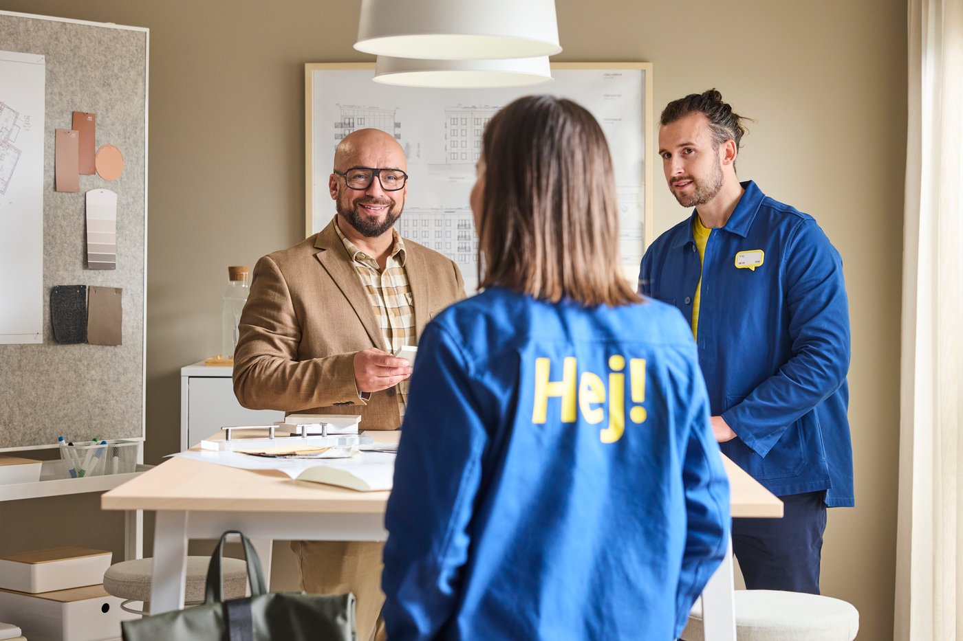 Two IKEA coworkers wearing blue Hej jackets are standing at a table talking to a customer who is smiling at them.