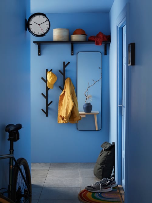 Hallway with yellow jacket and hat on TJUSIG hangers next to mirror, shelf and wall clock, with backpack next to door.