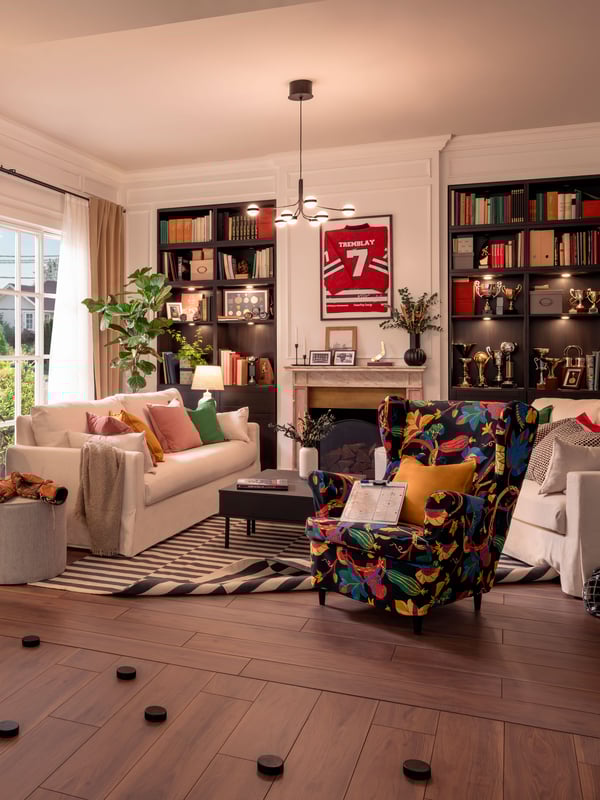 There is a large bookshelf filled with books against the back wall, flanked by two windows with white frames. A striking red poster with the number ‘7’ hangs above the bookshelf. The furniture includes a white sofa adorned with pastel-colored pillows, a black coffee table with white accents, and an armchair with a vibrant floral pattern. The floor is covered by dark wooden planks and a black-and-white striped area rug. Scattered on the floor are several small, round, black objects that resemble stones or decorative pieces. The room exudes warmth and appears to be an inviting space for relaxation or reading.