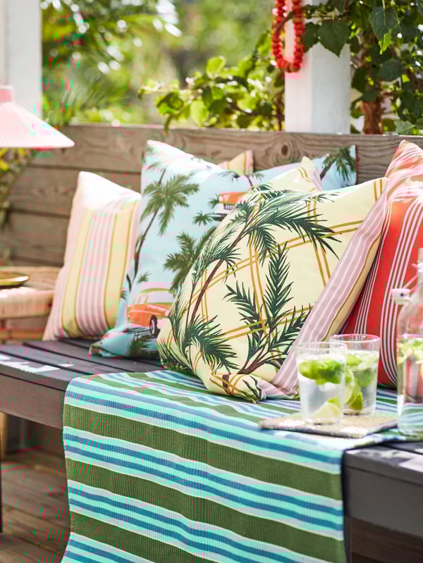 Outdoor setting with a wooden bench semi-filled with cushions with SOMMAR 2020 covers and two glasses on a small tray.