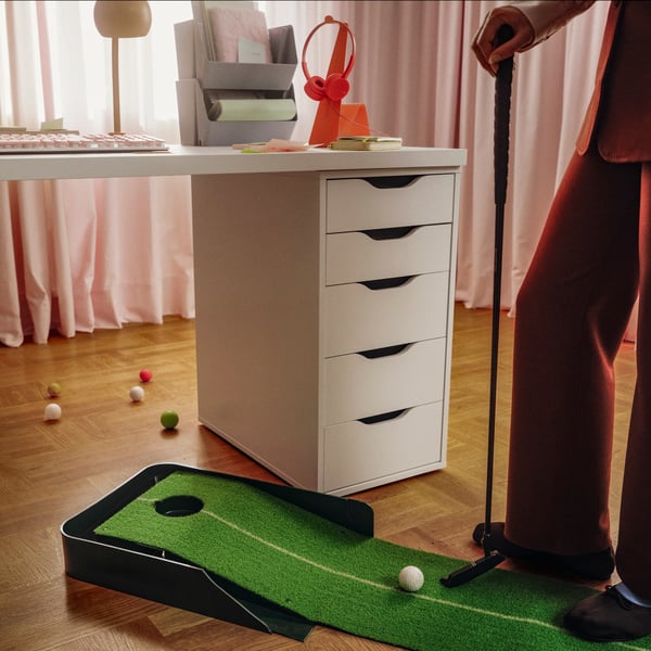 A white ALEX drawer unit with sleek, cut-out handles supports a white desk. Next to it, a person practices golf putts.
