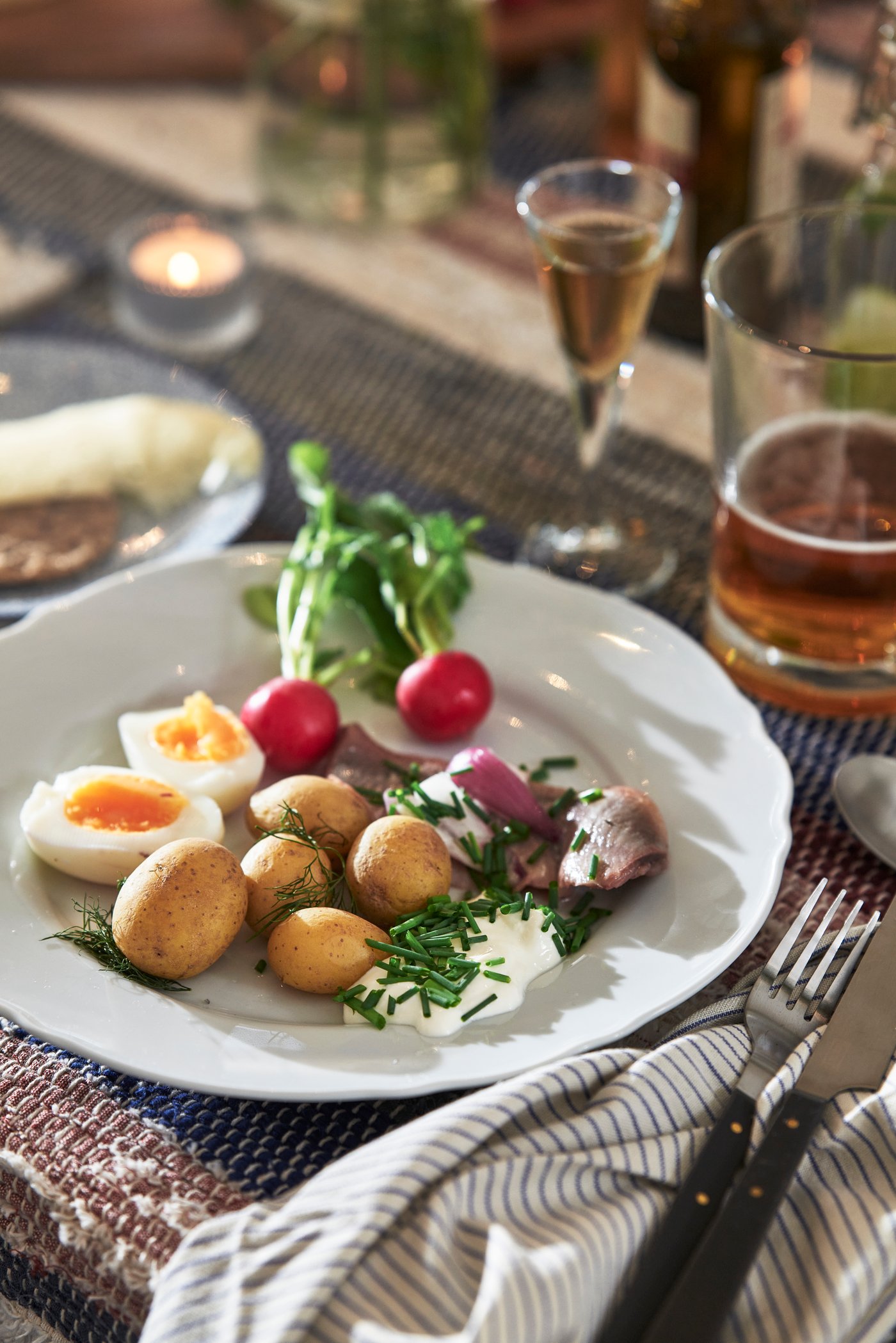 Teller mit Salzkartoffeln, halbierten Eiern, Radieschen, Kräutern und Hering, dazu Besteck auf gestreifter Stoffserviette und Tischläufer.