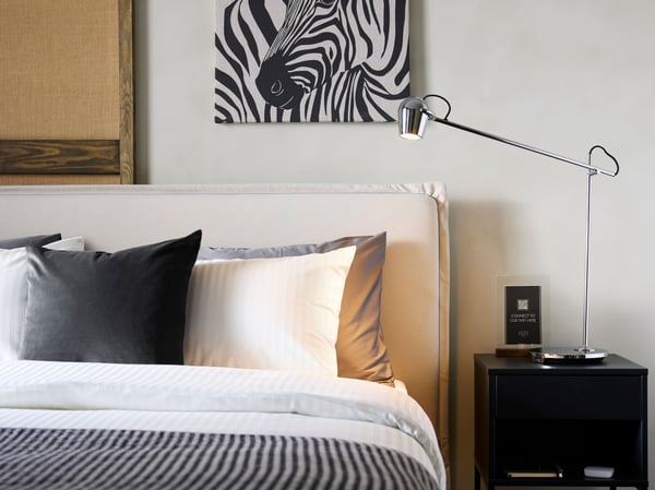 A beige TÄRNKULLEN upholstered bed frame accents a freshly made hotel bed on a wall below a zebra artwork, by a nightstand.