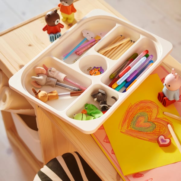 A white TROFAST tray with compartments holding pens and pencils sits on top of a TROFAST tabletop with a drawing beside it.