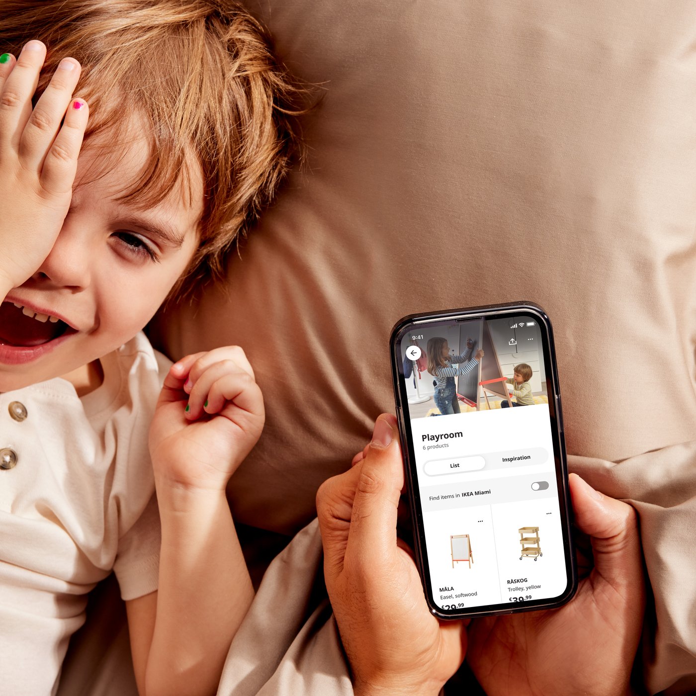 A child lies on a pillow beside an adult holding a smartphone that displays the IKEA app.