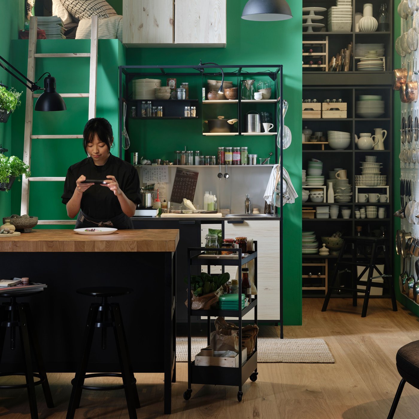 En person står ved en køkkenø og tager billeder af mad på en tallerken. Et ÄSPINGE minikøkken foran en grøn væg.