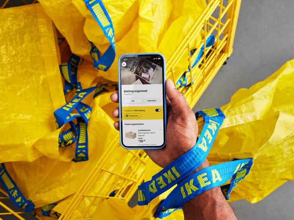 A hand holds a smartphone displaying the IKEA app, surrounded by yellow IKEA bags with blue handles in a shopping cart.