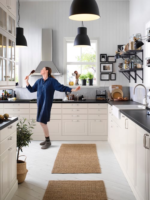 Une cuisine moderne dans laquelle se trouvent des armoires inférieures avec tiroirs, une personne, une hotte murale, une vitrine et des tapis. 