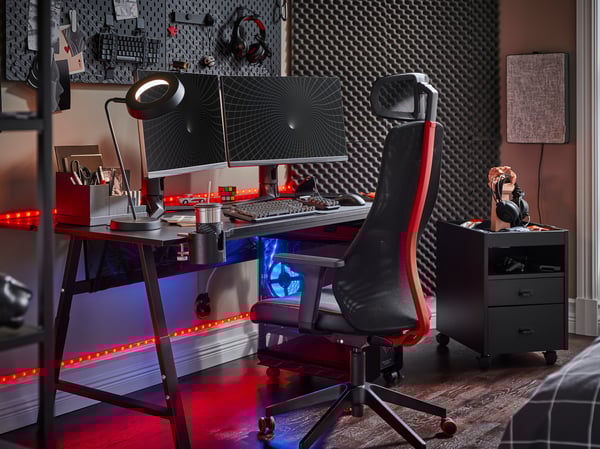 A black and red MATCHSPEL gaming chair faces two screens on a black UTESPELARE gaming desk and a wall of gaming accessories.
