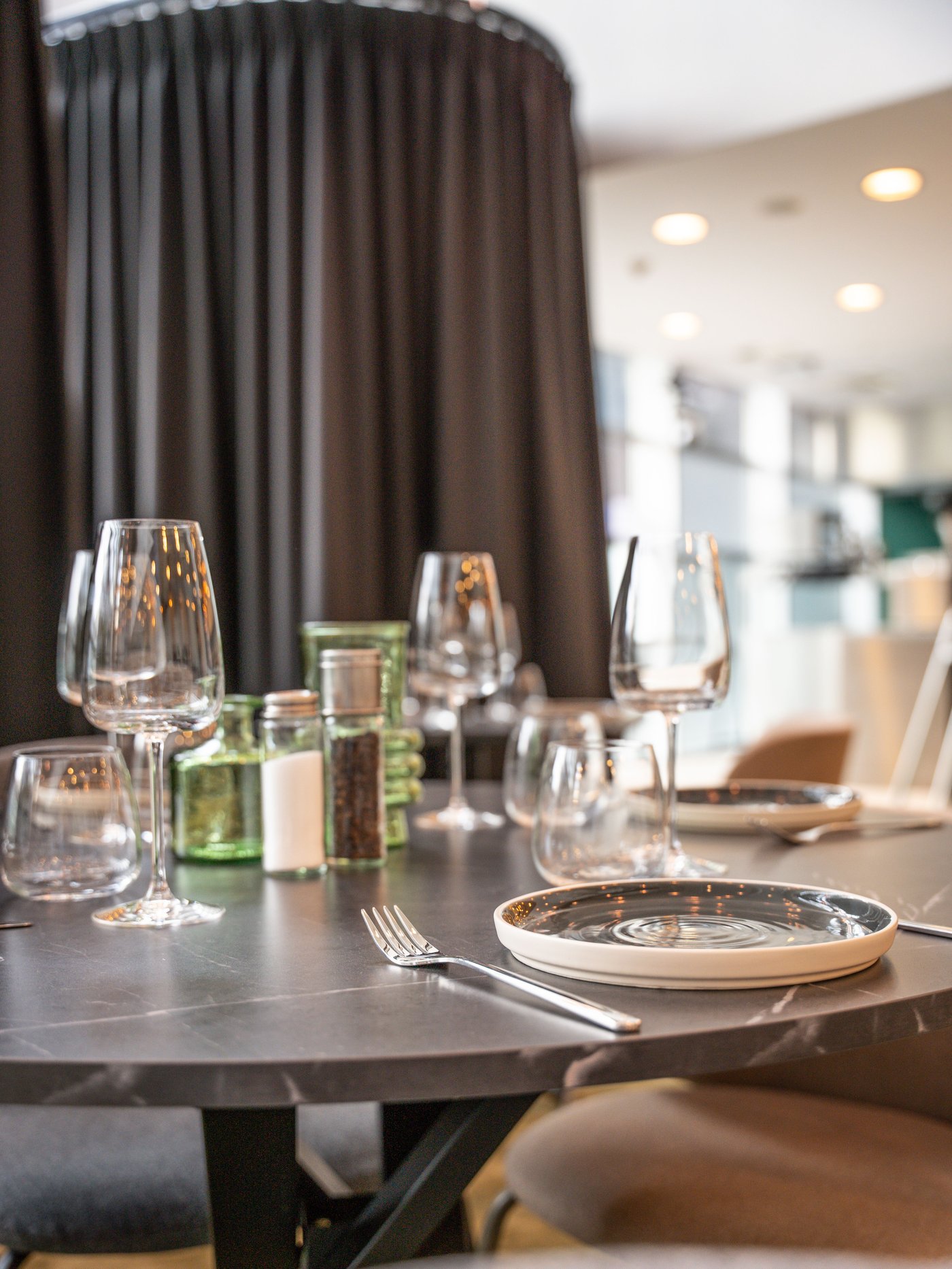 Elegante restauranttafel met glazen, borden en groene potjes op donker blad. Achteraan gordijnen en sfeervolle verlichting in modern interieur.