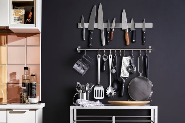 Kitchen wall with magnetic knife rack and hanging utensils above a counter with cookware and bottles.
