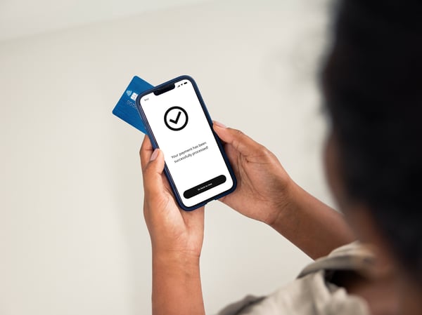 A person holds a smartphone showing a green checkmark on the screen, with a blue credit card partially visible behind it.