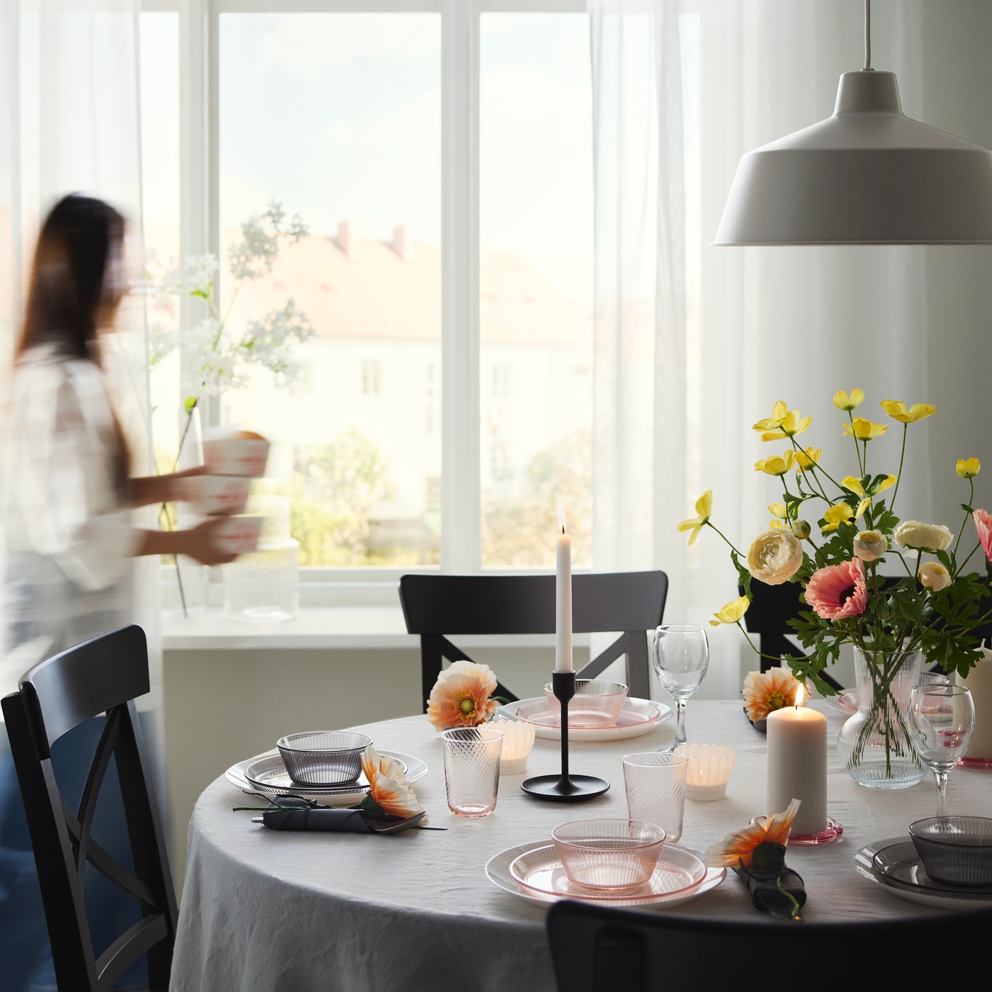 A dining table with a tablecloth and black ROSENTORP chairs is set for a meal with dinnerware, glasses, flowers and candles.