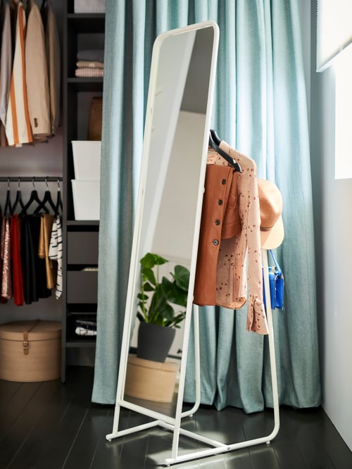 Ein KNAPPER Standspiegel, auf dessen Rückseite Kleidung und eine Handtasche hängen, steht vor einem Vorhang, der einen Kleiderschrank verdeckt.