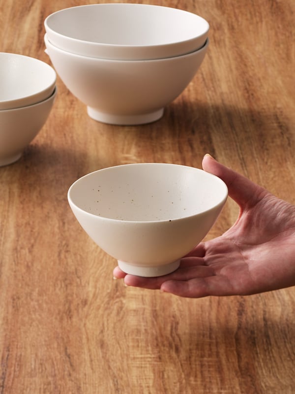 木製テーブルの上で、手に持たれたシンプルな白い陶器の茶碗。奥には同じ茶碗が重ねて置かれている。
