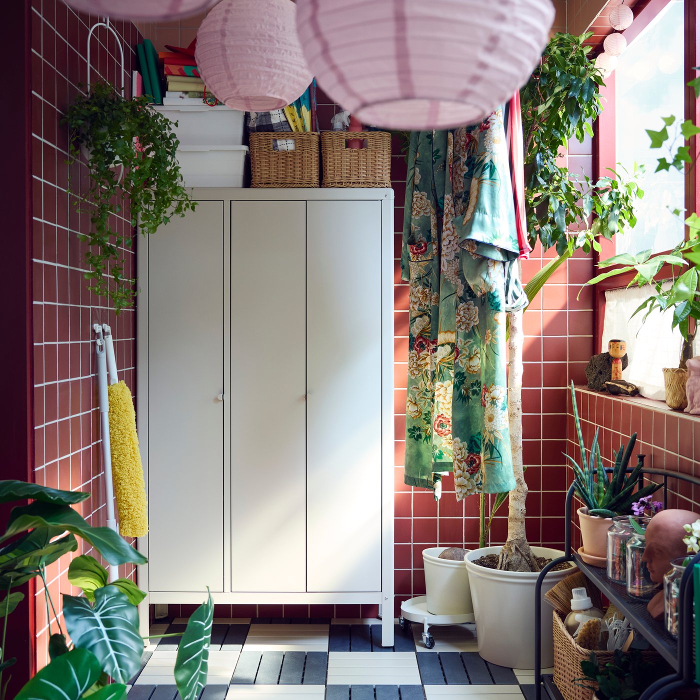 A balcony with a beige KOLBJÖRN cabinet made for outdoor use, surrounded by hanging plants, clothes and pink lamps.