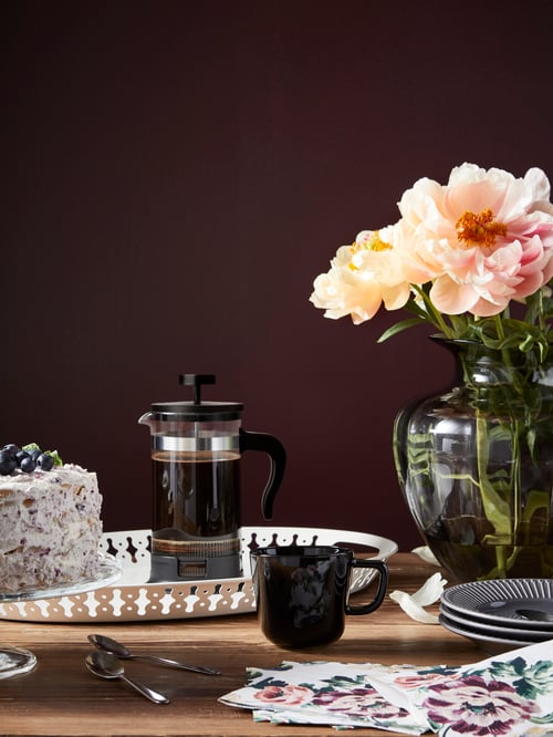 A coffee maker and cake placed on a tray with a mug, paper napkin, vase and cutlery placed alongside.