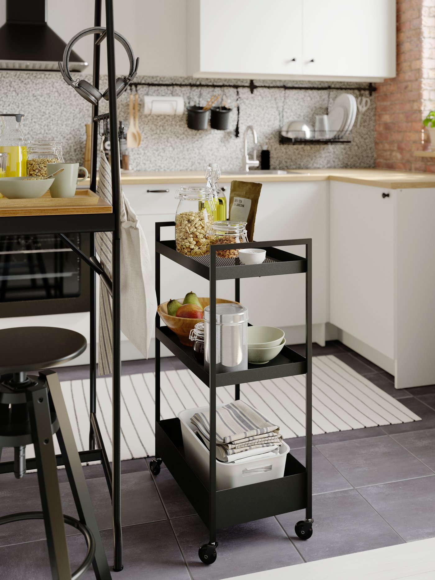 Una pequeña cocina blanca con un carrito NISSAFORS negro junto a una mesa alta con cereales, frutas, cuencos y una caja de paños de cocina.