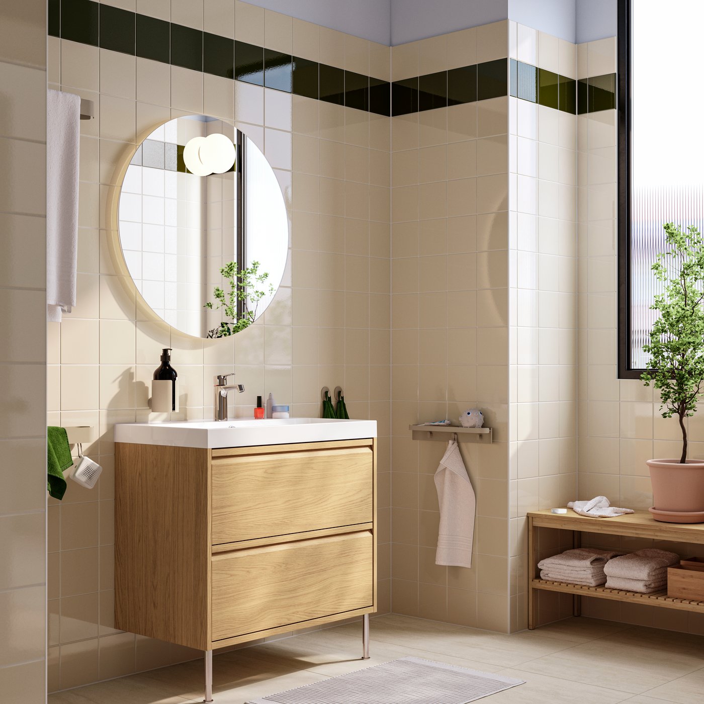 Sotto uno specchio circolare c'è un mobile lavabo ÄNGSJÖN/BACKSJÖN effetto rovere con due cassetti e un lavabo, su due gambe di metallo.