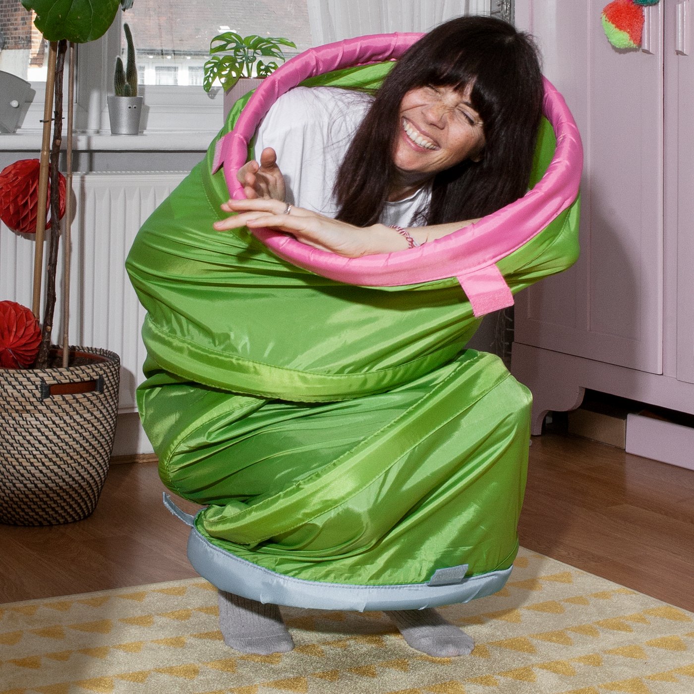 A laughing woman has crept into a BUSA play tunnel and half sits, half stands on the carpet in a children’s room.