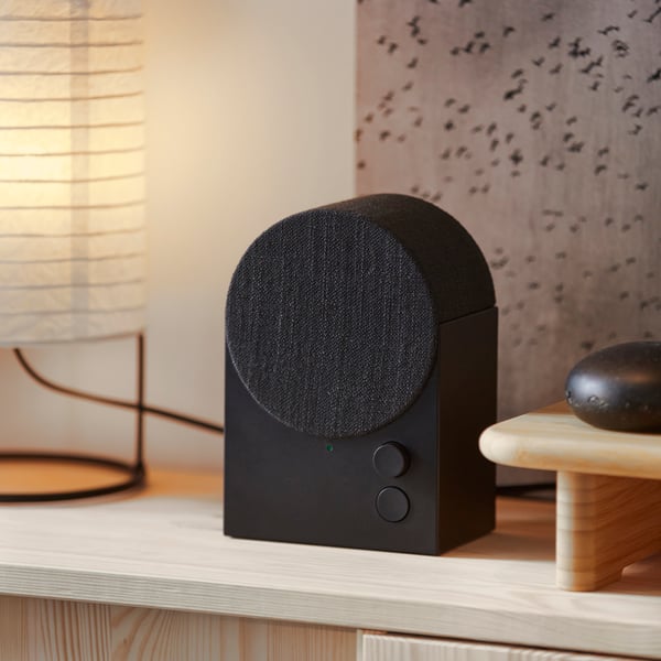 The black NATTBAD Bluetooth speaker with a rounded shape and two buttons on the front is standing on top of a pine cabinet.