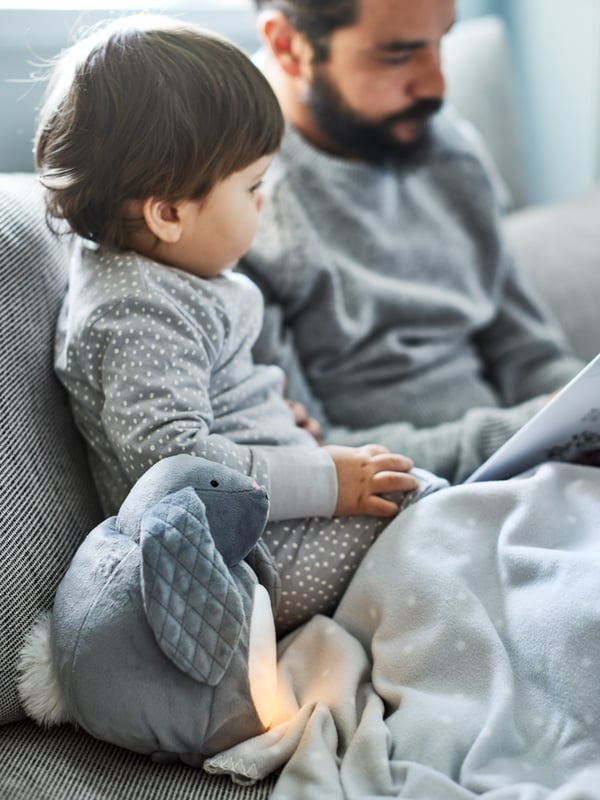 A man reads a children’s book to a young child sitting on a sofa beside a PEKHULT soft toy with LED night light.