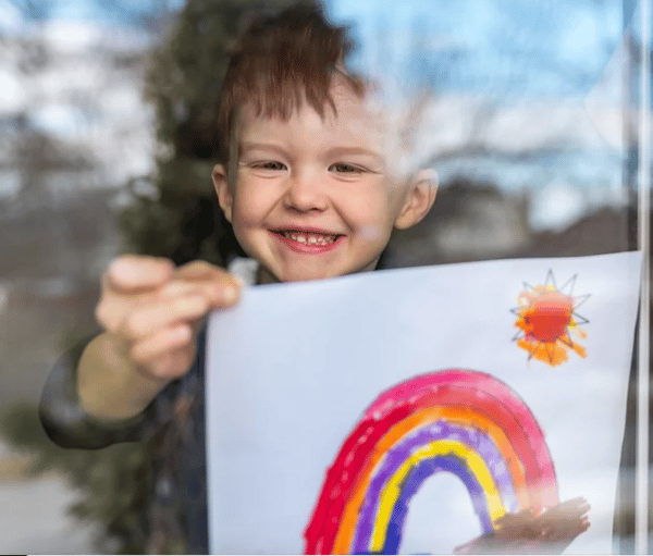 Kind toont een tekening van een regenboog en zon achter een raam.