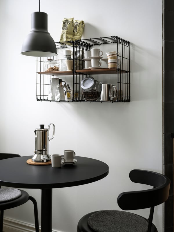 Two GULLHULT wall shelves holding coffee and tea accessories hangs above a small, round table and two chairs.