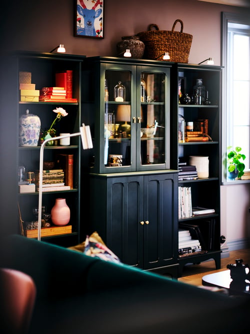 Une armoire LOMMARP bleu-vert foncé avec des portes vitrées flanquées de deux étagères LOMMARP, toutes remplies de livres et d’ornements.